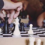 Ein Foto von einer weißen Hand, die eine schwarze Schahfigur auf einem Schachbrett zieht. Auf dem Schachbrett sind verschiedene weiße und schwarze Figuren zu sehen. Auch am Rande des Brettes liegen ein paar Figuren.