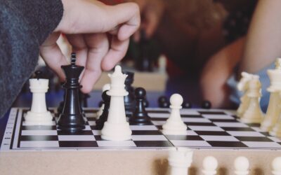 Ein Foto von einer weißen Hand, die eine schwarze Schahfigur auf einem Schachbrett zieht. Auf dem Schachbrett sind verschiedene weiße und schwarze Figuren zu sehen. Auch am Rande des Brettes liegen ein paar Figuren.