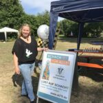 Laura Schalkhäuser ist eine mittelgroße Frau mit langen blonden Haaren. Sie trägt ein schwarzes T-Shirt und eine hellblaue Jeans. Sie steht neben einem blauem Stand. Zwischen dem Stand und ihr steht eine Werbewand, die ein Plakat zeigt. Auf dem Plakat ist der Schriftzug Schach ist für alle zu lesen und das logo vom Schachklub Arnstein zu sehen. Laura Schalkhäuser lächelt.