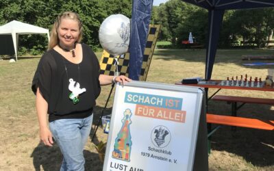 Laura Schalkhäuser ist eine mittelgroße Frau mit langen blonden Haaren. Sie trägt ein schwarzes T-Shirt und eine hellblaue Jeans. Sie steht neben einem blauem Stand. Zwischen dem Stand und ihr steht eine Werbewand, die ein Plakat zeigt. Auf dem Plakat ist der Schriftzug Schach ist für alle zu lesen und das logo vom Schachklub Arnstein zu sehen. Laura Schalkhäuser lächelt.