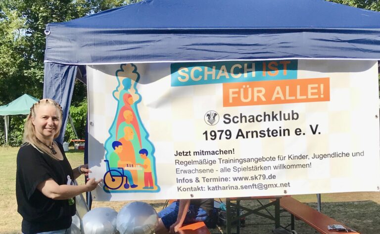 Laura Schalkhäuser ist eine mittelgroße Frau mit langen blonden Haaren. Sie steht neben einem blauem Stand. Vor dem Stand ist ein Banner gespannt, auf dem der Schriftzug Schach ist für alle steht. Laura Schalkhäuser lächelt und deutet auf den Banner mit einem Finger hin.