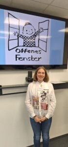 Das Bild zeigt Tatiana Flores beim Mädchenschachkongress 2025 in Frankfurt. Tatiana Flores eist eine mittelgroße, blonde, junge Frau, die vor einem Bildschirm lächelnd, steht. Auf dem Bilschirm ist das Logo vom Offenen Fenster auf einem blauen Hintergrund zu sehen.