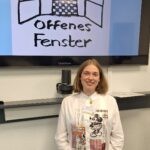 Das Bild zeigt Tatiana Flores beim Mädchenschachkongress 2025 in Frankfurt. Tatiana Flores eist eine mittelgroße, blonde, junge Frau, die vor einem Bildschirm lächelnd, steht. Auf dem Bilschirm ist das Logo vom Offenen Fenster auf einem blauen Hintergrund zu sehen.