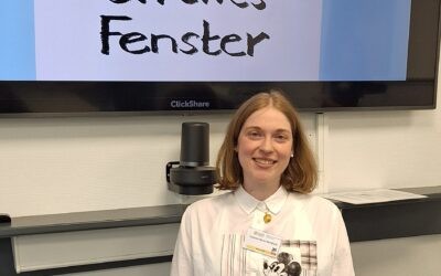 Das Bild zeigt Tatiana Flores beim Mädchenschachkongress 2025 in Frankfurt. Tatiana Flores eist eine mittelgroße, blonde, junge Frau, die vor einem Bildschirm lächelnd, steht. Auf dem Bilschirm ist das Logo vom Offenen Fenster auf einem blauen Hintergrund zu sehen.