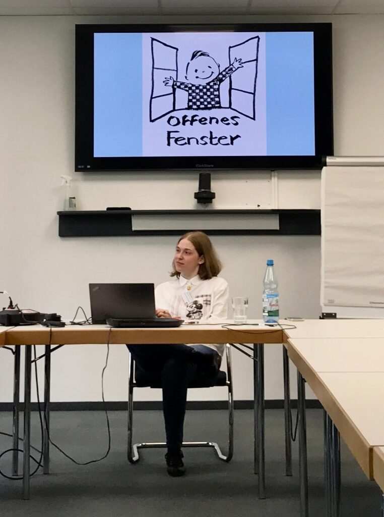 Das Bild zeigt Tatiana Flores beim Mädchenschachkongress 2025 in Frankfurt. Tatiana Flores eist eine mittelgroße, blonde, junge Frau, die vor einem Bildschirm sitzt und einen Vortrag hält. Auf dem Bilschirm ist das Logo vom Offenen Fenster auf einem blauen Hintergrund zu sehen.