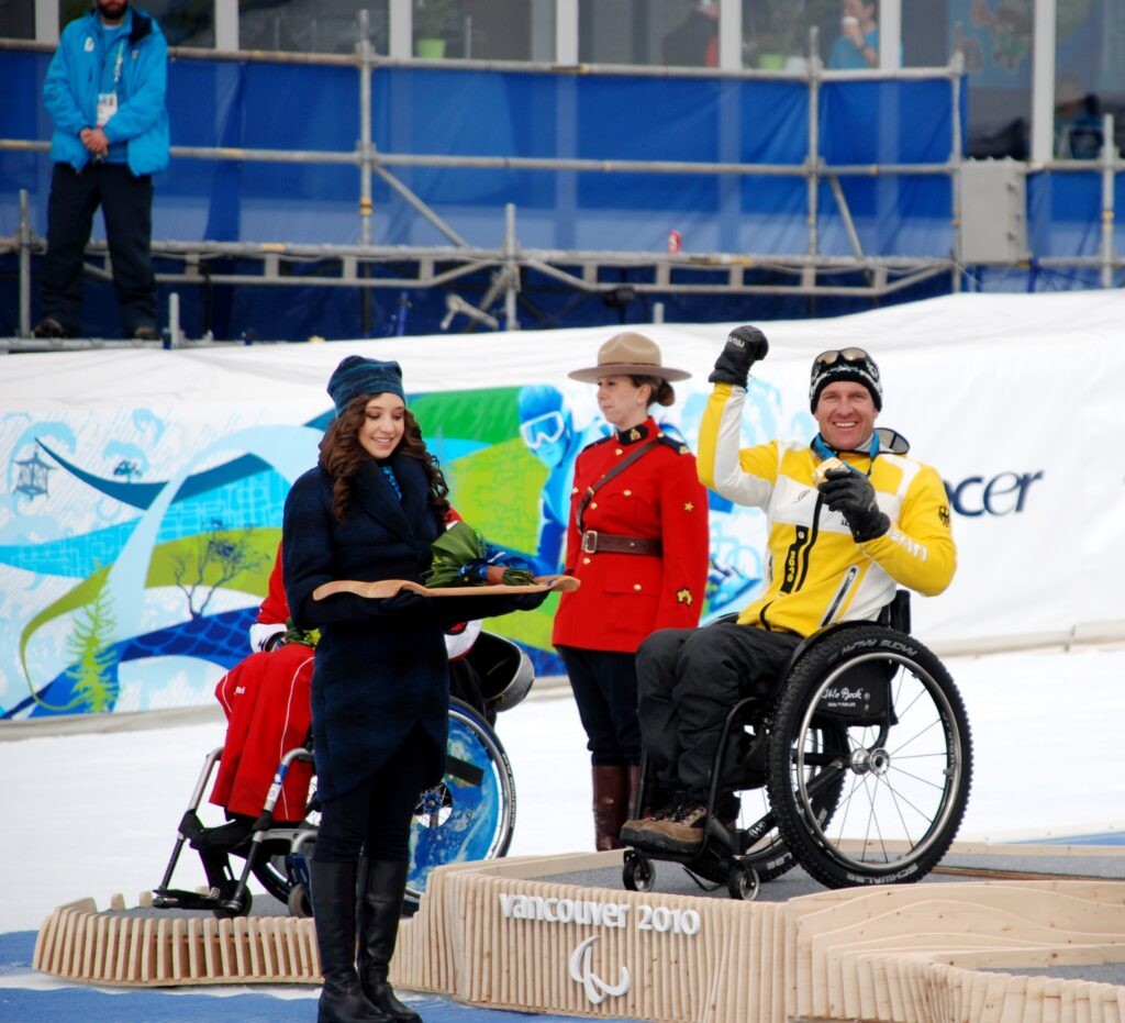 Ein Foto von Martin Braxenthaler bei der Medaillen-Übergabe von den Paralympics in Vancouver in 2010. Martin Braxenthaler sitzt in einem Rollstuhl auf einem Podium, ist mit warmer Winterkleidung angezogen. Er lächelt in die Kamera und hält eine goldene Medaille hoch.